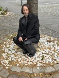 Karl Heinz Hirschberger crouching by a tree in a black blazer and leather trousers on a cobblestone sidewalk.