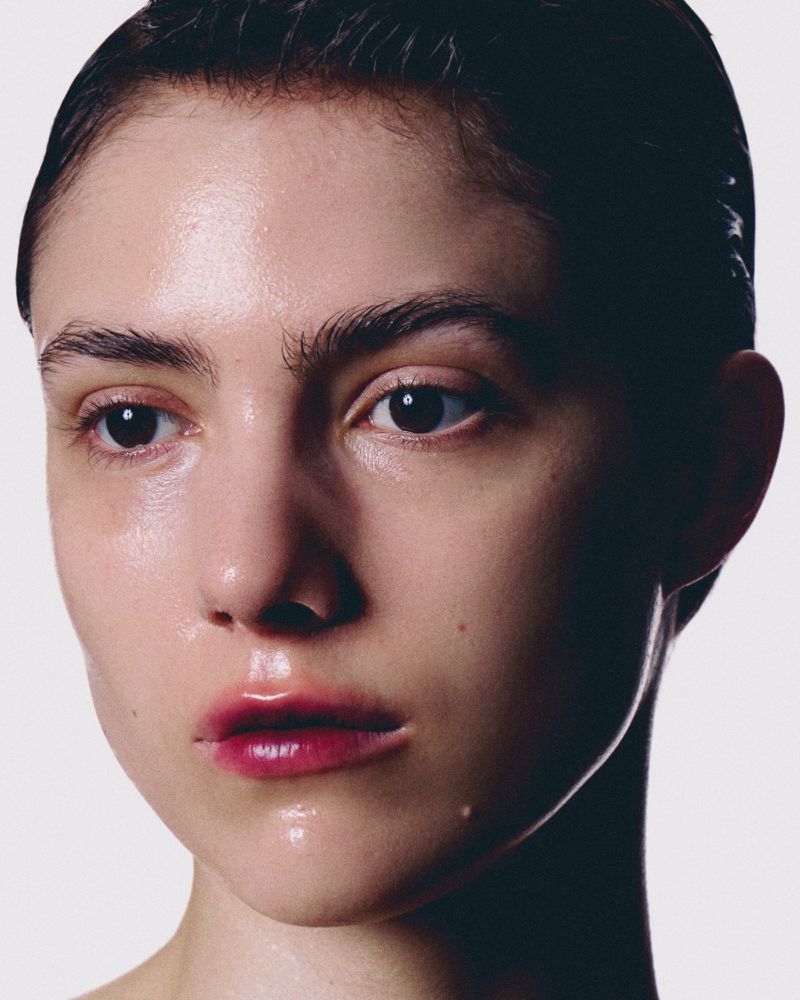 Close-up portrait of a woman with wet, slicked-back hair and dewy skin against a plain white background.