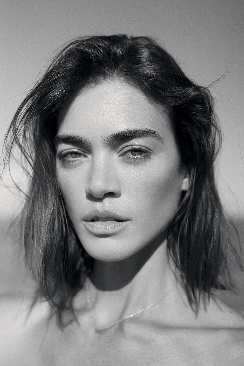 Black and white close-up portrait of a woman with tousled dark hair and defined eyebrows wearing a delicate chain necklace.