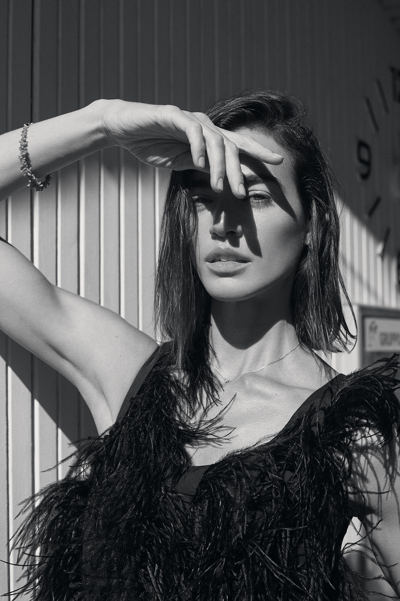 A woman with dark hair posing in a black feathered top, with her hand shielding her face from bright sunlight, against a paneled wall.