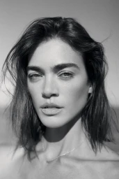 Black and white close-up portrait of a woman with tousled dark hair and defined eyebrows wearing a delicate chain necklace.