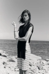Monochrome fashion portrait of a woman standing on a rocky beach wearing a draped black top and patterned shorts.