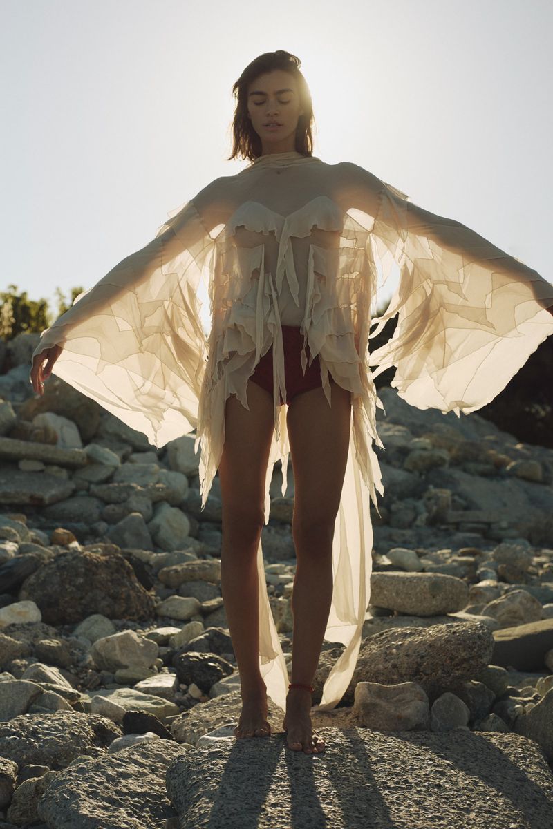 Model in a sheer off-white winged cape garment standing barefoot on rocks backlit by sunlight.
