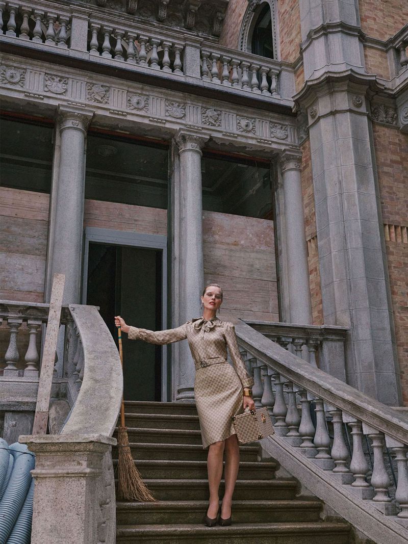 Eva Herzigova in a patterned beige silk skirt suit holding a broom and handbag on a grand stone staircase.