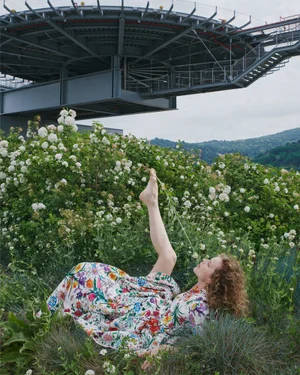 Eva Herzigova lying among green foliage and white flowers in a floral dress with one leg raised beneath a steel structure.
