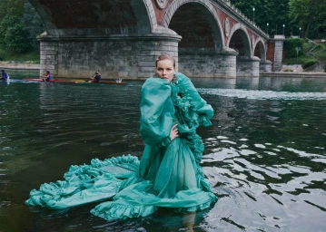 Eva Herzigova standing in a river wearing a voluminous ruffled green gown in front of a classical arched stone bridge.