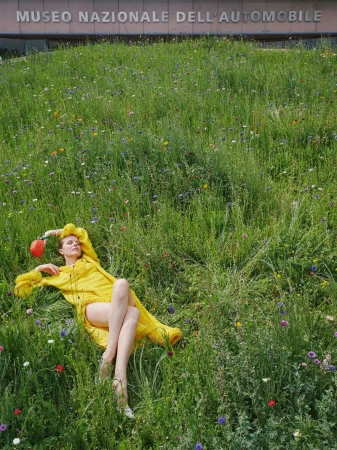 Model Eva Herzigova reclines on a lush grassy slope among wildflowers wearing a bright yellow knit dress and holding a small red accessory.