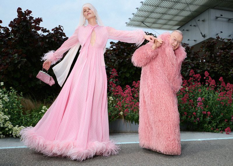 Eva Herzigova in a pink feather-trimmed gown posing alongside Walter Pfeiffer in an oversized pink shaggy coat outdoors.