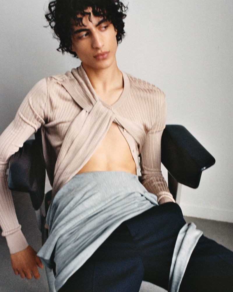 A young adult male with dark curly hair sits on a black chair wearing a beige long sleeve top with a gray garment over navy pants.