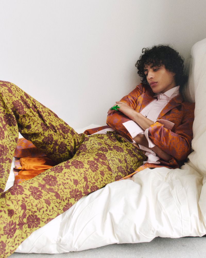 A young man with curly hair lounges on a white bed, wearing a patterned orange blazer and floral pants in a relaxed pose.