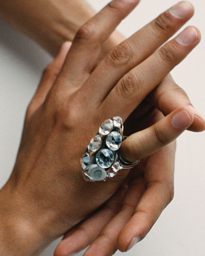 Close-up on a hand wearing a silver ring encrusted with multiple clear gemstones, set against a plain white background.