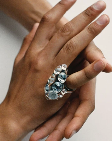 Close-up on a hand wearing a silver ring encrusted with multiple clear gemstones, set against a plain white background.