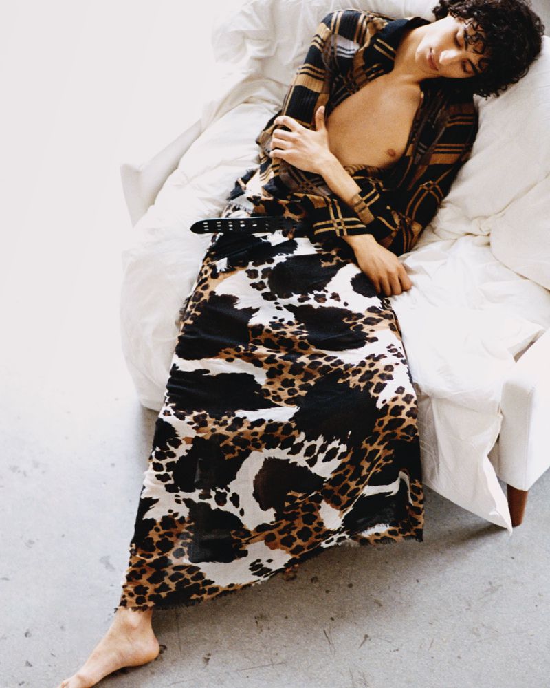 Man in patterned shirt and skirt lounges in bed. Model wears brown and black animal print skirt, black and brown checkered top, relaxed pose.