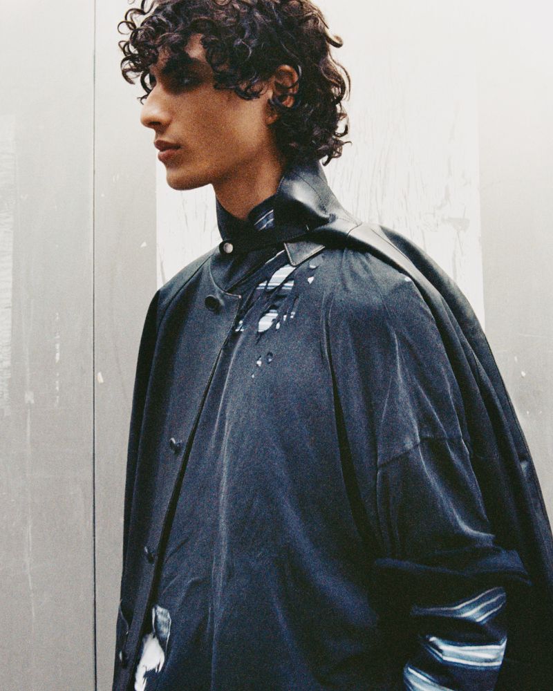 Profile of a young man with curly dark hair wearing a layered distressed dark jacket against a neutral grey backdrop.