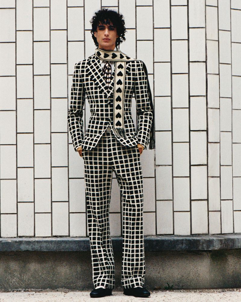 Adult male poses in a black and white checkered suit with a black heart scarf against a white tile wall, fashion shoot.