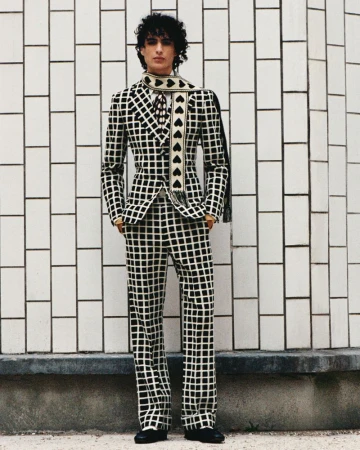 Adult male poses in a black and white checkered suit with a black heart scarf against a white tile wall, fashion shoot.
