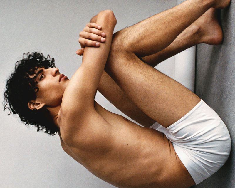 Young adult man with curly dark hair, wearing white underwear, posing shirtless against a solid light grey background in a studio.