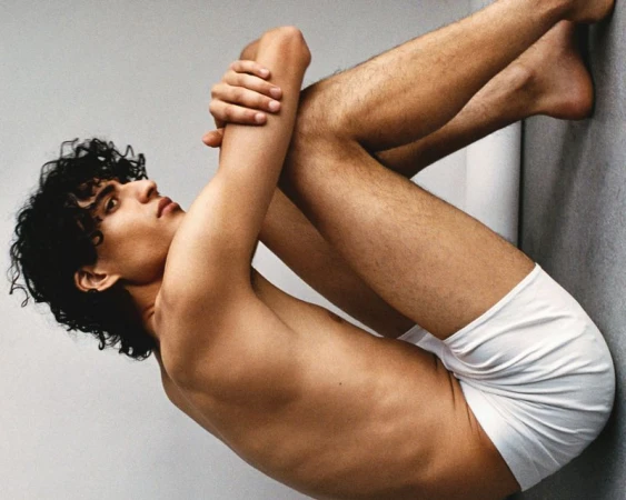 Young adult man with curly dark hair, wearing white underwear, posing shirtless against a solid light grey background in a studio.