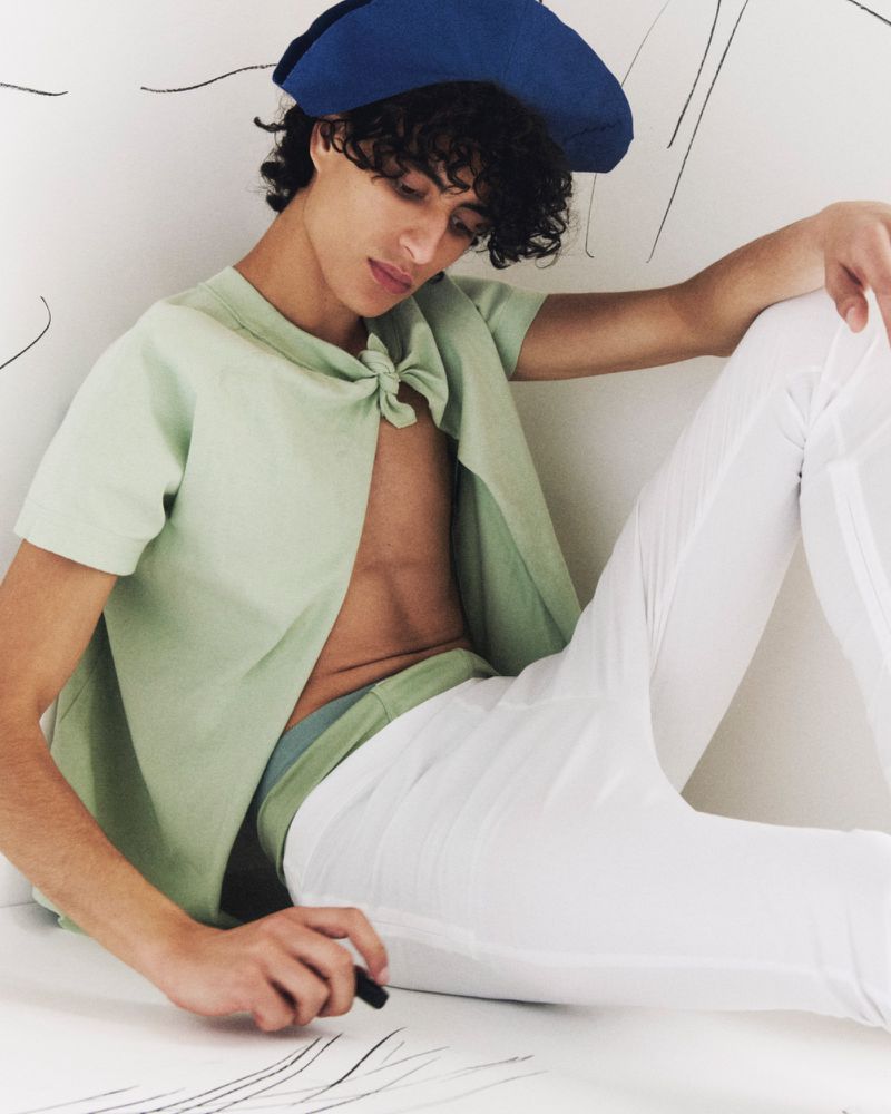 Young adult male with curly hair in unbuttoned green top and white pants sits drawing on paper wearing a blue cap indoors.