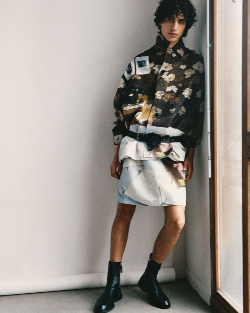 Adult male fashion model poses in jacket, light blue skirt, and black boots in a studio. White walls and natural light.