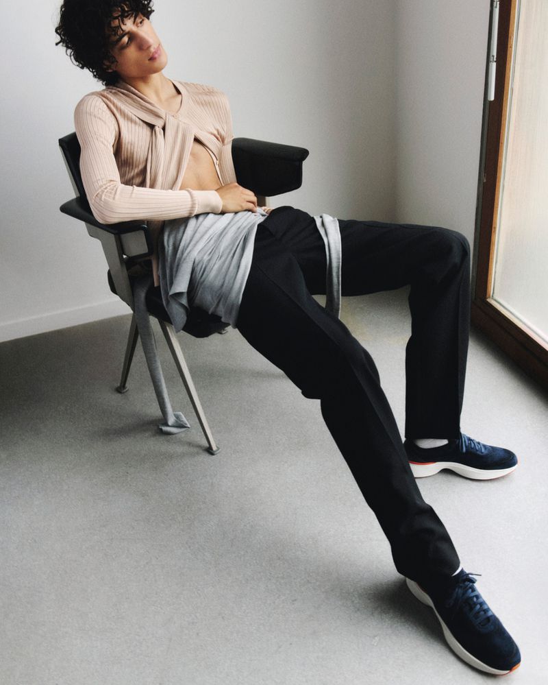 Young adult with curly hair is sitting on a chair in pants, sneakers, and a twisted top in a bright indoor setting near a window.