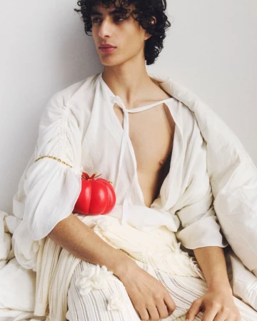 Portrait of a young adult with curly hair wearing a white outfit, seated indoors, holding a bright red tomato-shaped object in a studio setting.
