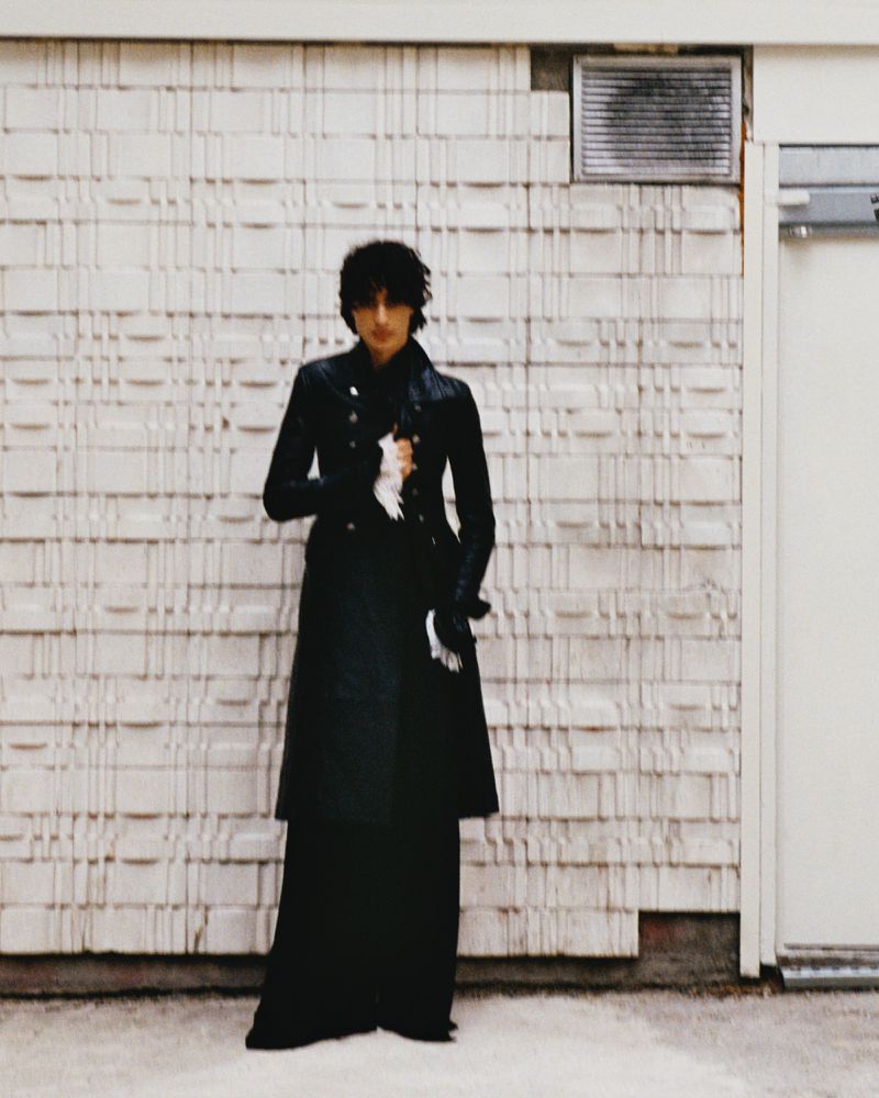 An adult wears a long black coat and stands in front of a textured beige wall next to a white door with a dark gray vent above.