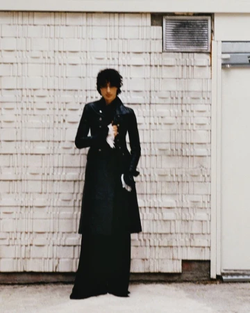 An adult wears a long black coat and stands in front of a textured beige wall next to a white door with a dark gray vent above.