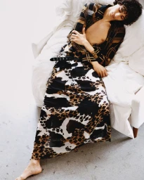 Model Yoesry Detre reclines on a white sofa wearing a patterned skirt and open plaid shirt for NSS, photographed by Joan Braun.