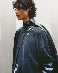 Profile of a young man with curly dark hair wearing a layered distressed dark jacket against a neutral grey backdrop.