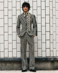 Model Yoesry Detre posing in a black and white grid patterned suit and heart print scarf against a white tiled wall.