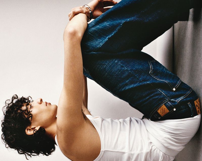 Model with curly dark hair wearing a white tank top and blue denim jeans posing against a neutral studio backdrop.