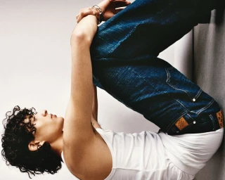 Model with curly dark hair wearing a white tank top and blue denim jeans posing against a neutral studio backdrop.