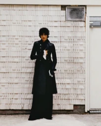 Model Yoesry Detre in a long black tailored coat and wide-leg trousers standing against a textured relief wall.