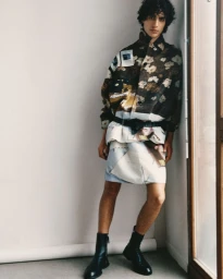 Model Yoesry Detre posing in a printed collage jacket, printed skirt, and black leather boots against a white wall.