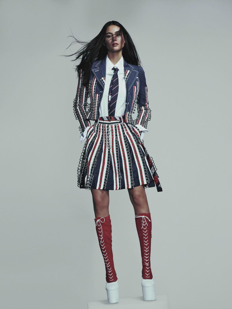Model posing in a navy and red striped jacket and pleated skirt with red lace-up knee-high boots against a grey studio backdrop.