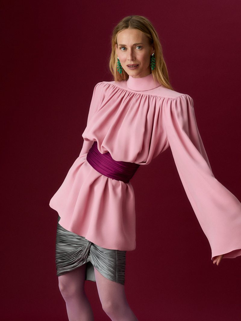 Woman in pink high-neck long-sleeve top with purple wide waist belt and shimmering grey skirt against a deep burgundy studio background.