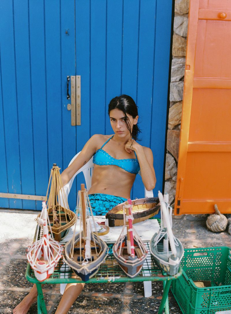 Model in a bright turquoise bikini seated behind a table of handcrafted model boats in front of a blue door.