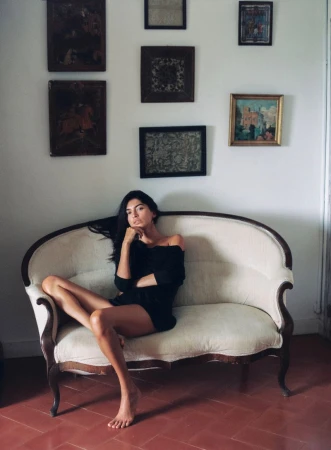 Woman with long dark hair wearing a black off-the-shoulder dress seated barefoot on a vintage cream sofa beneath framed wall pictures.