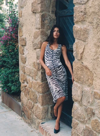 Model wearing a black and white zebra-print midi dress leaning in a stone doorway next to flowering greenery.