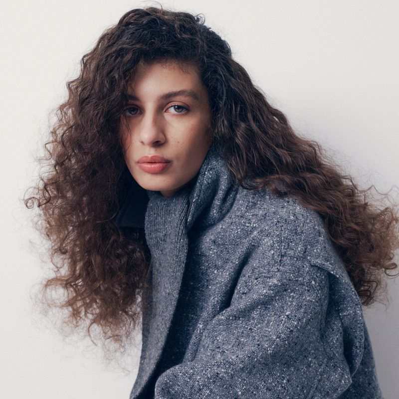 Model with voluminous curly hair wearing a textured grey wool coat with a high ribbed collar against a white studio background.
