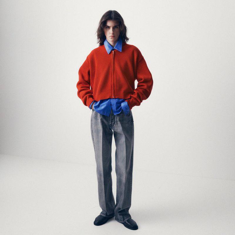 A male model standing against a white backdrop, wearing a bright orange knit zip-up jacket over a blue shirt and grey tailored trousers.