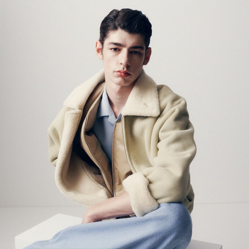 Male model seated in a cream fleece jacket and light blue trousers against a plain white studio background.