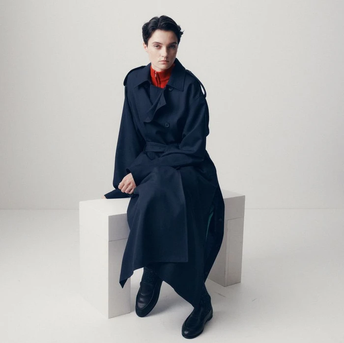 Model seated on a white block wearing an oversized navy trench coat and red high-neck top against a white studio background.