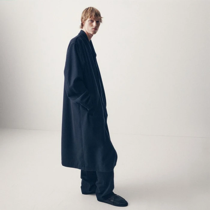 Model wearing a long dark navy overcoat and matching wide trousers against a white studio background.