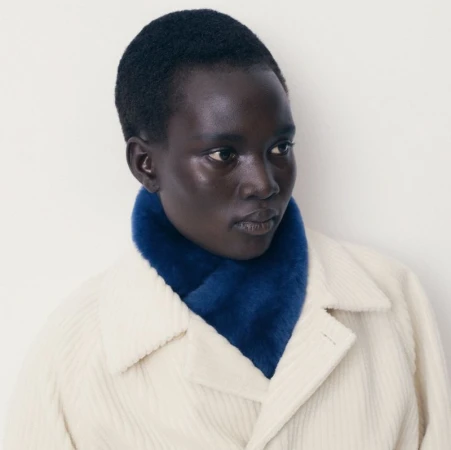 Close-up of a model wearing a deep blue plush neck warmer and a cream ribbed coat against a plain off-white studio background.