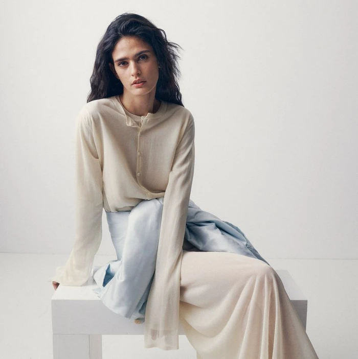 Woman seated on a white block wearing a lightweight cream long-sleeve top with a pale blue shirt tied around her waist.