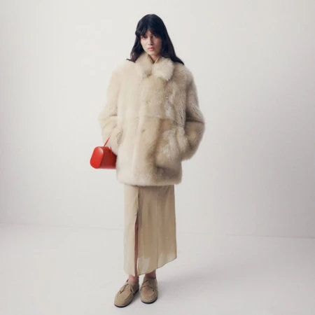 Model wearing an oversized cream textured coat and sheer beige skirt holding a bright red handbag against a seamless white background.