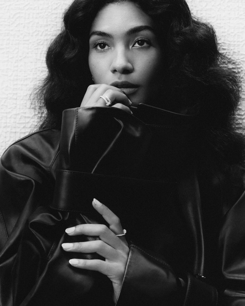 Black and white portrait of a woman with long dark wavy hair wearing a black leather jacket and silver rings posing in a studio.