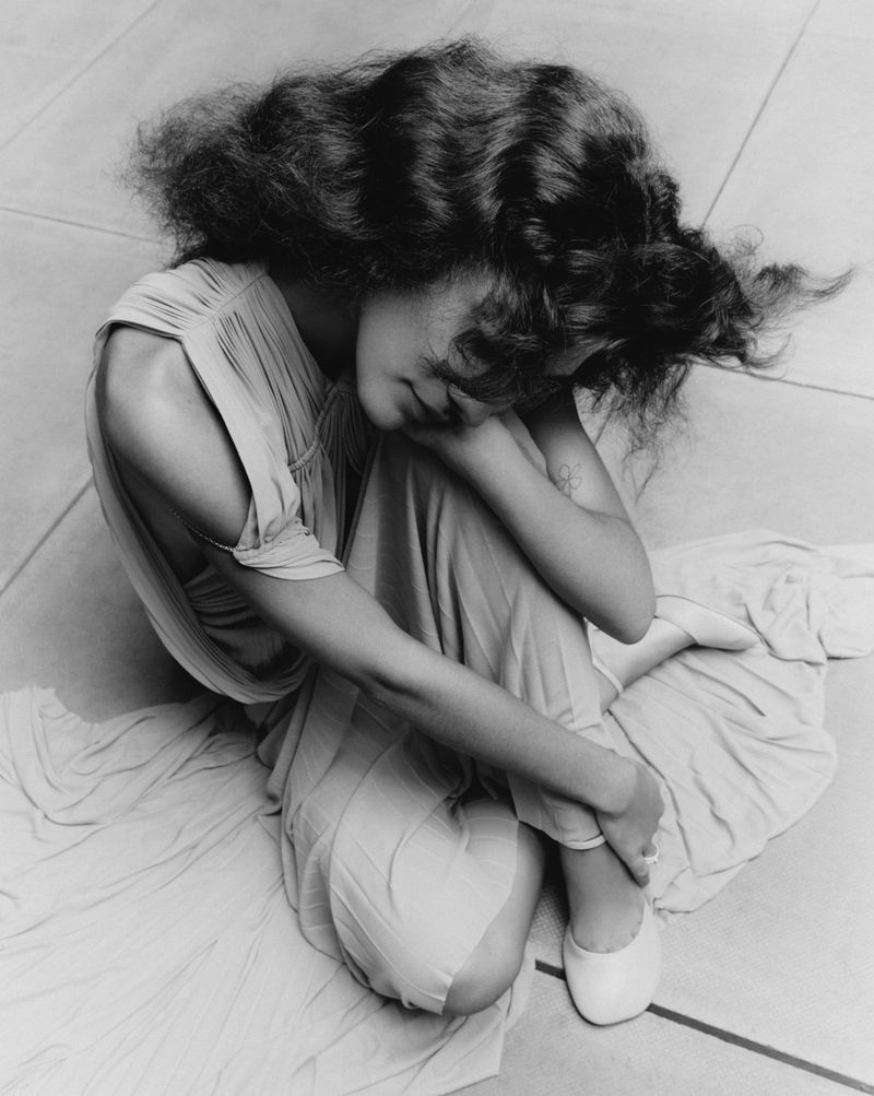 Black and white portrait of a woman with voluminous dark hair wearing a gown and ballet shoes, seated indoors with arms crossed.